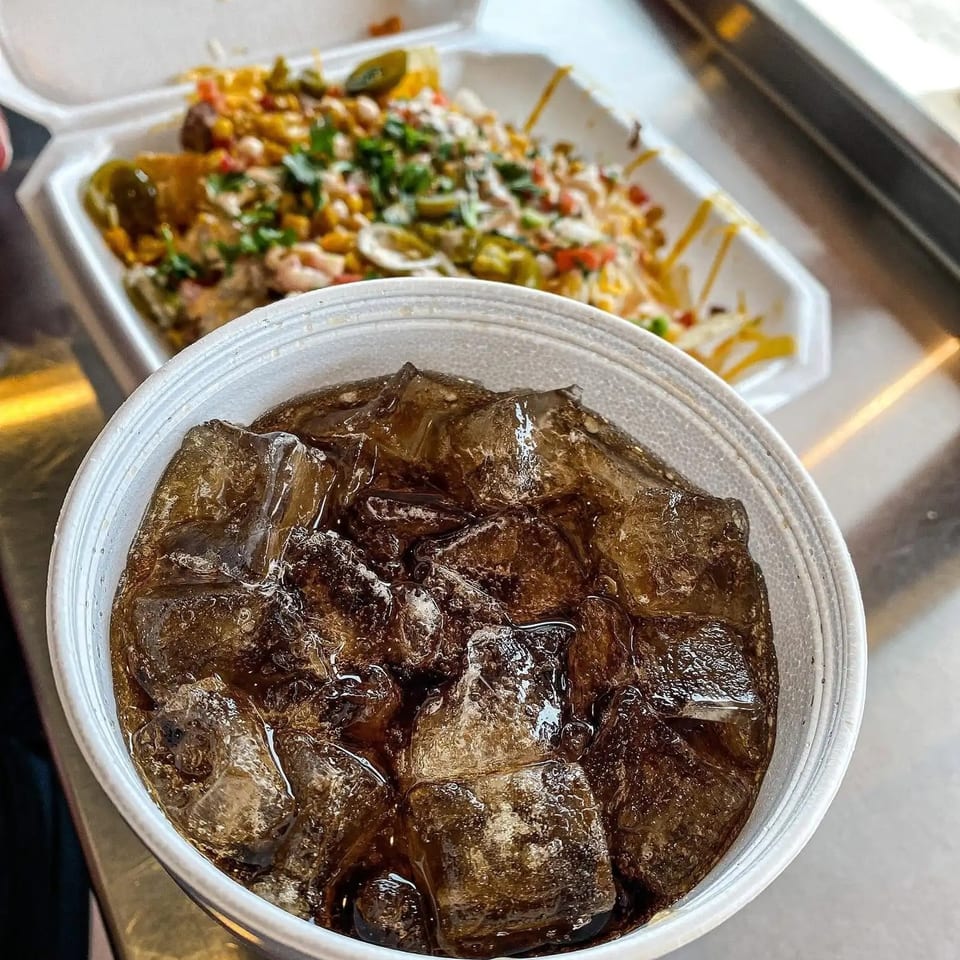Iced soda in a white cup, with a takeout container of loaded nachos in the background.