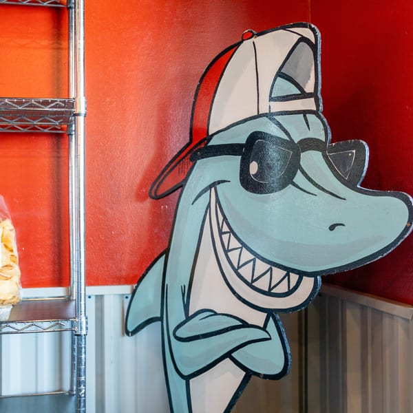 Large cartoon shark mural beside a metal shelving unit holding tortilla chips and condiment containers in a restaurant.