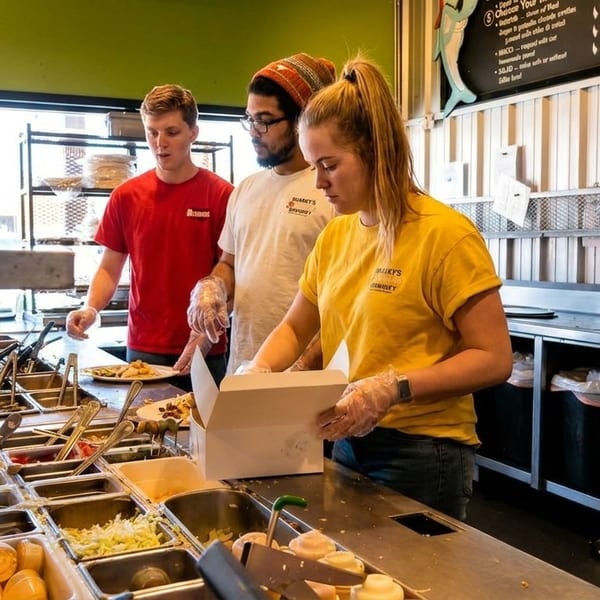 Three restaurant workers in branded shirts preparing food at a busy serving counter with many ingredients.