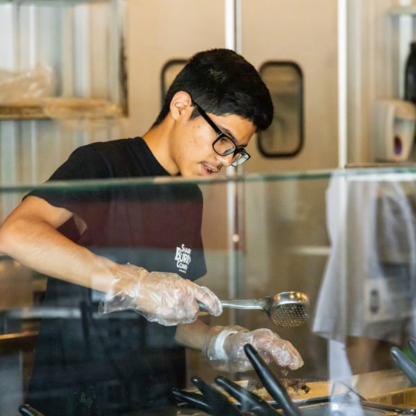 A young man in glasses and gloves serves food from a buffet line at a restaurant.